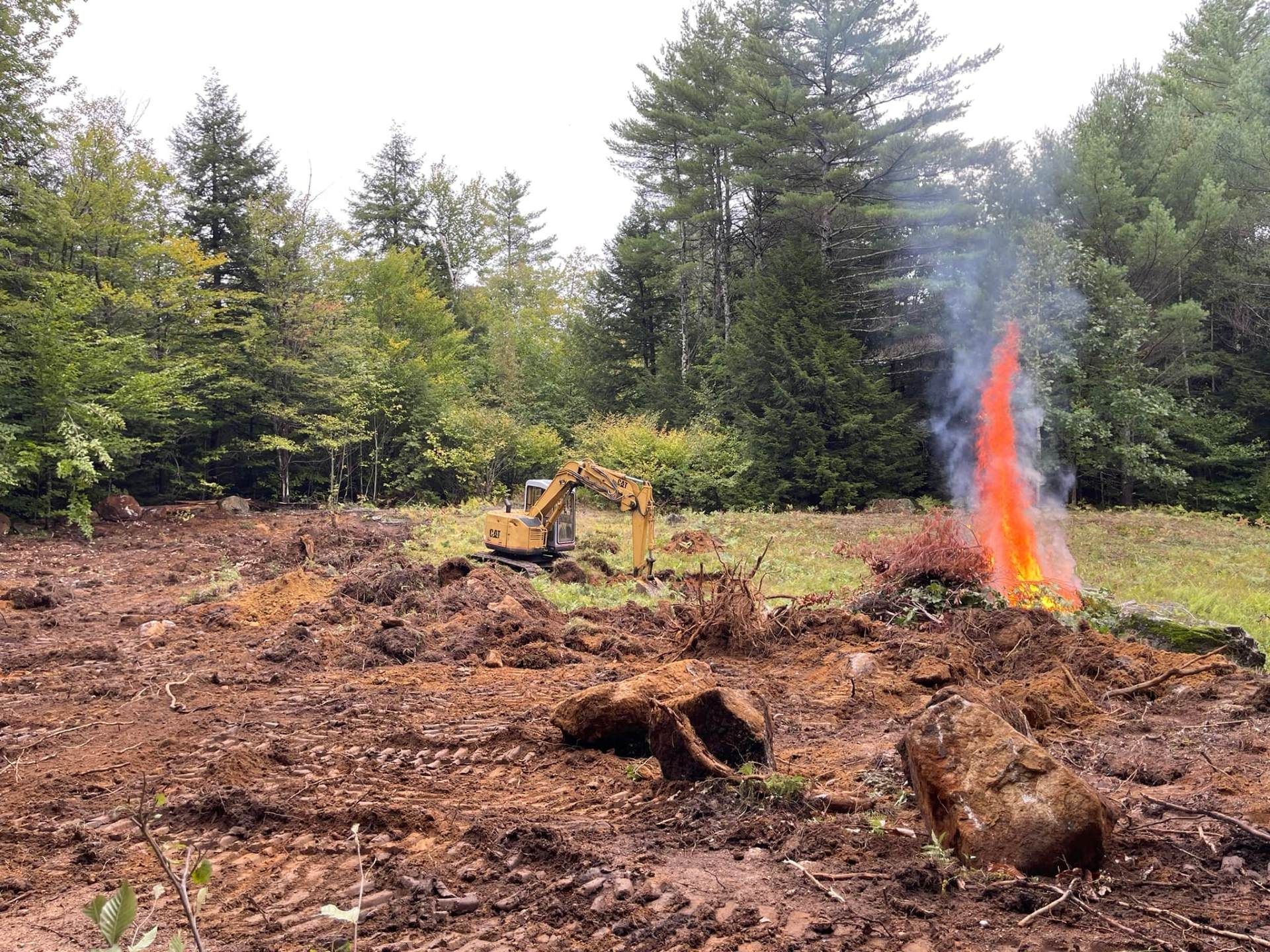 An excavator clearing land next to a burning brush pile in a wooded area.