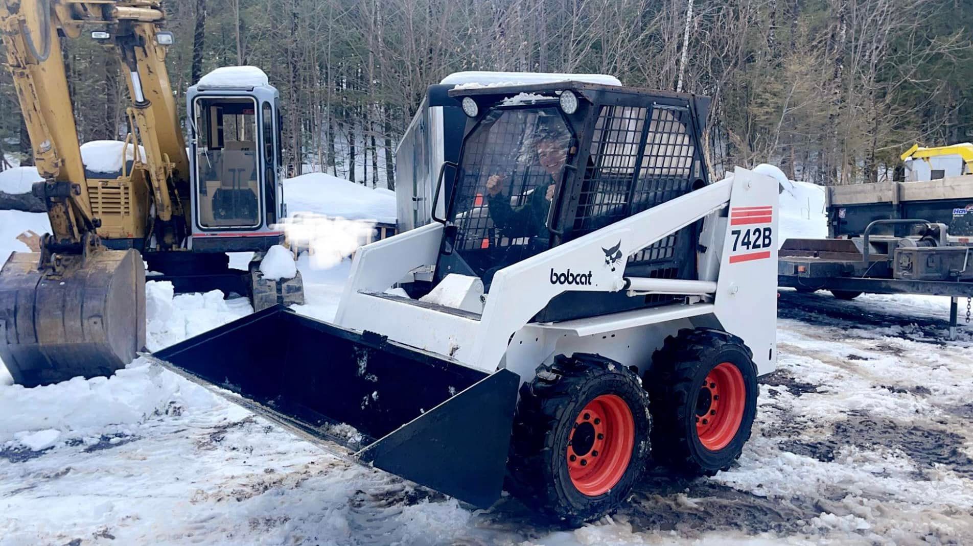 A white Bobcat 743B skid-steer loader with a front bucket, parked in the snow. An excavator is to the left.
