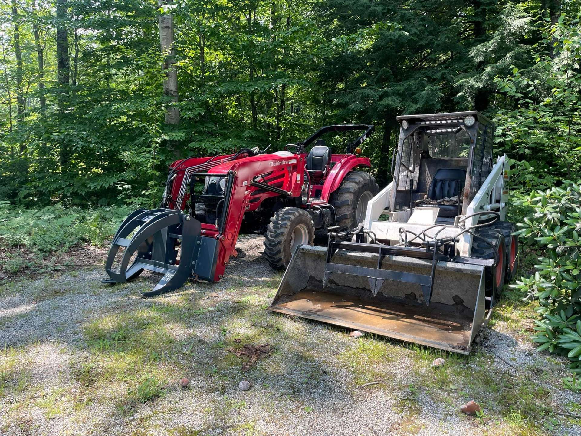 Red tractor with grapple attachment and a Bobcat skid-steer loader parked on grass near trees.