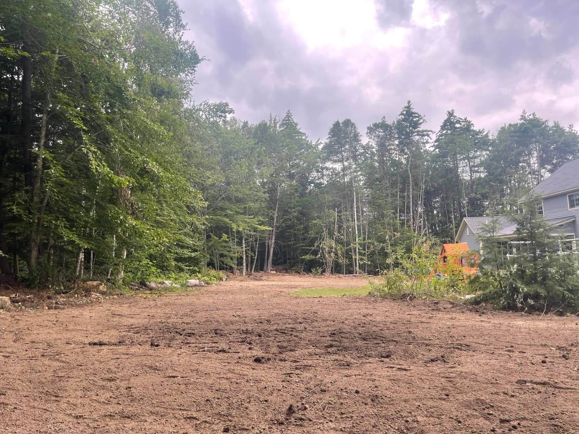 Cleared land with brown soil, surrounded by trees under a cloudy sky.