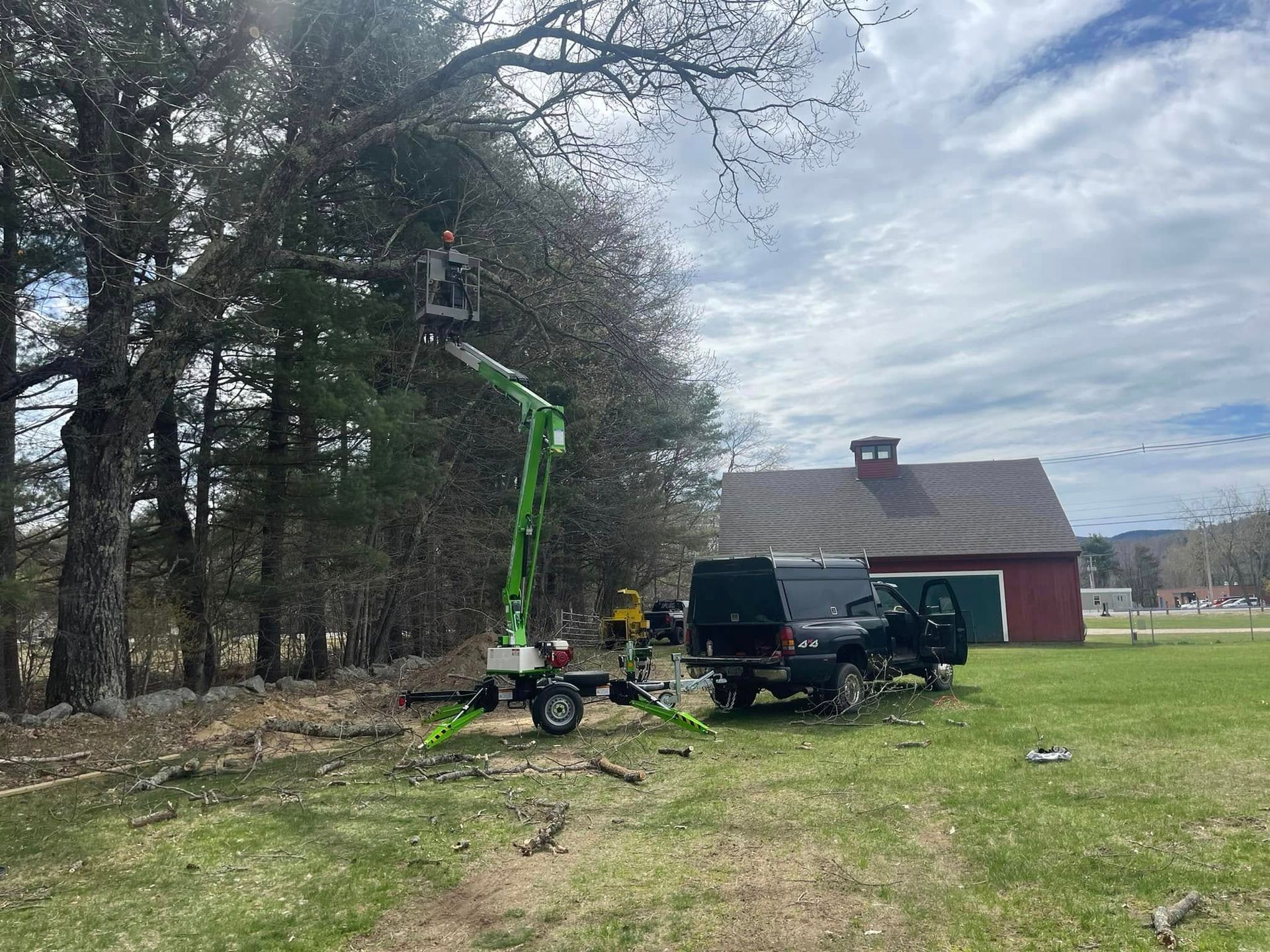 Man in lift trimming tree branches near a barn; a truck and trailer are present.