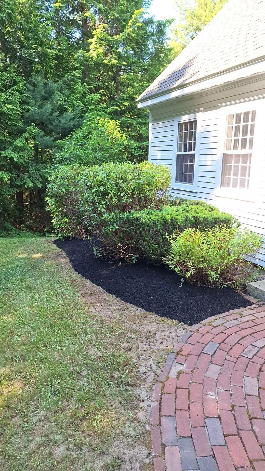 A house with white siding and windows, beside a shrub bed with fresh black mulch, and a brick pathway.