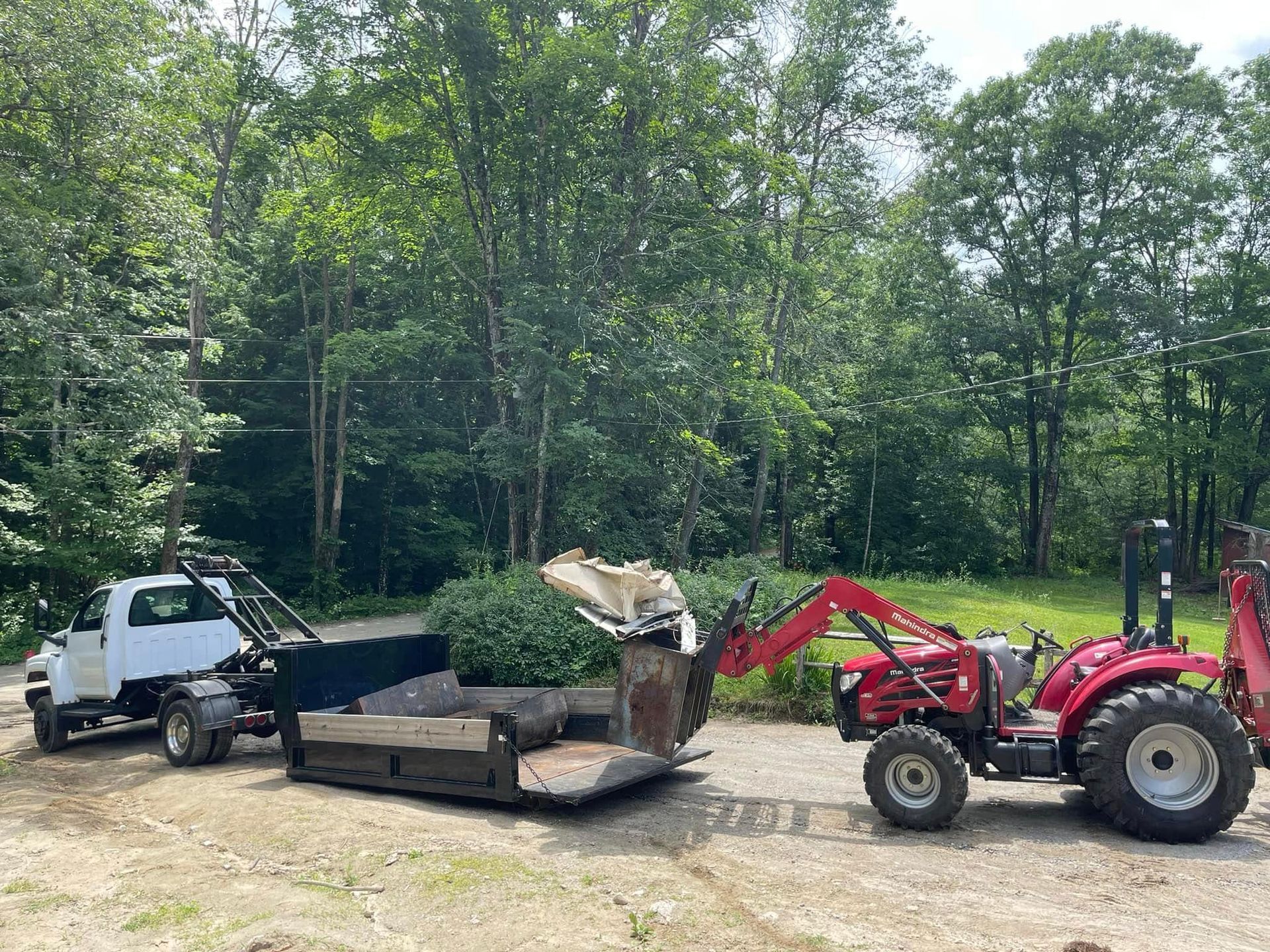 Red tractor using a front-end loader to dump debris into a trailer. A white truck is parked nearby. Outdoors, sunny setting.