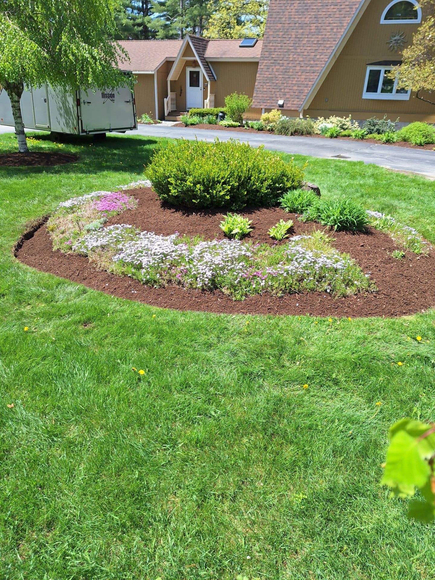 A circular garden bed with various plants and brown mulch, in front of a house.