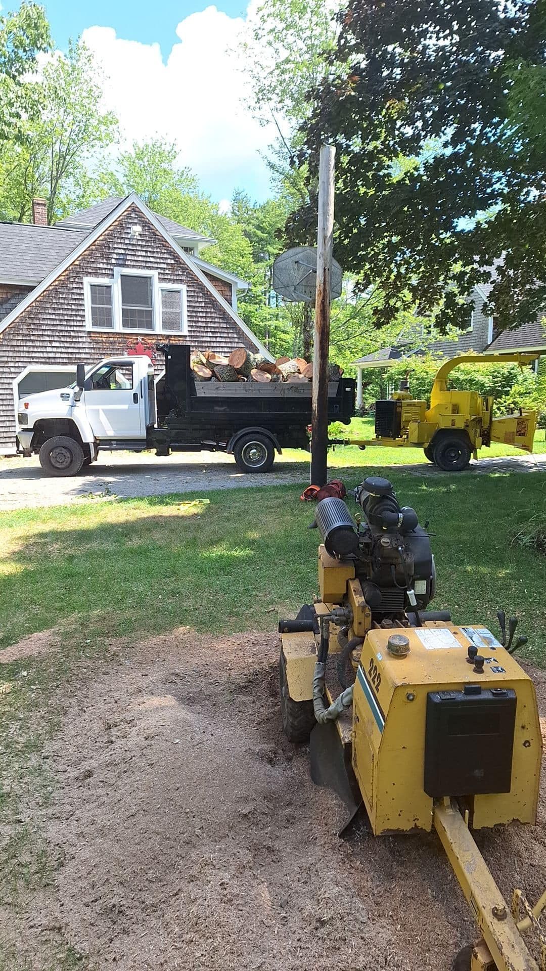 A stump grinder, truck, and chipper in a yard, near a house with logs in the truck bed, and trees.
