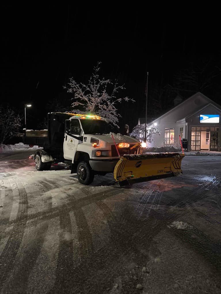 Snowplow truck clearing a snowy parking lot at night; building in the background.