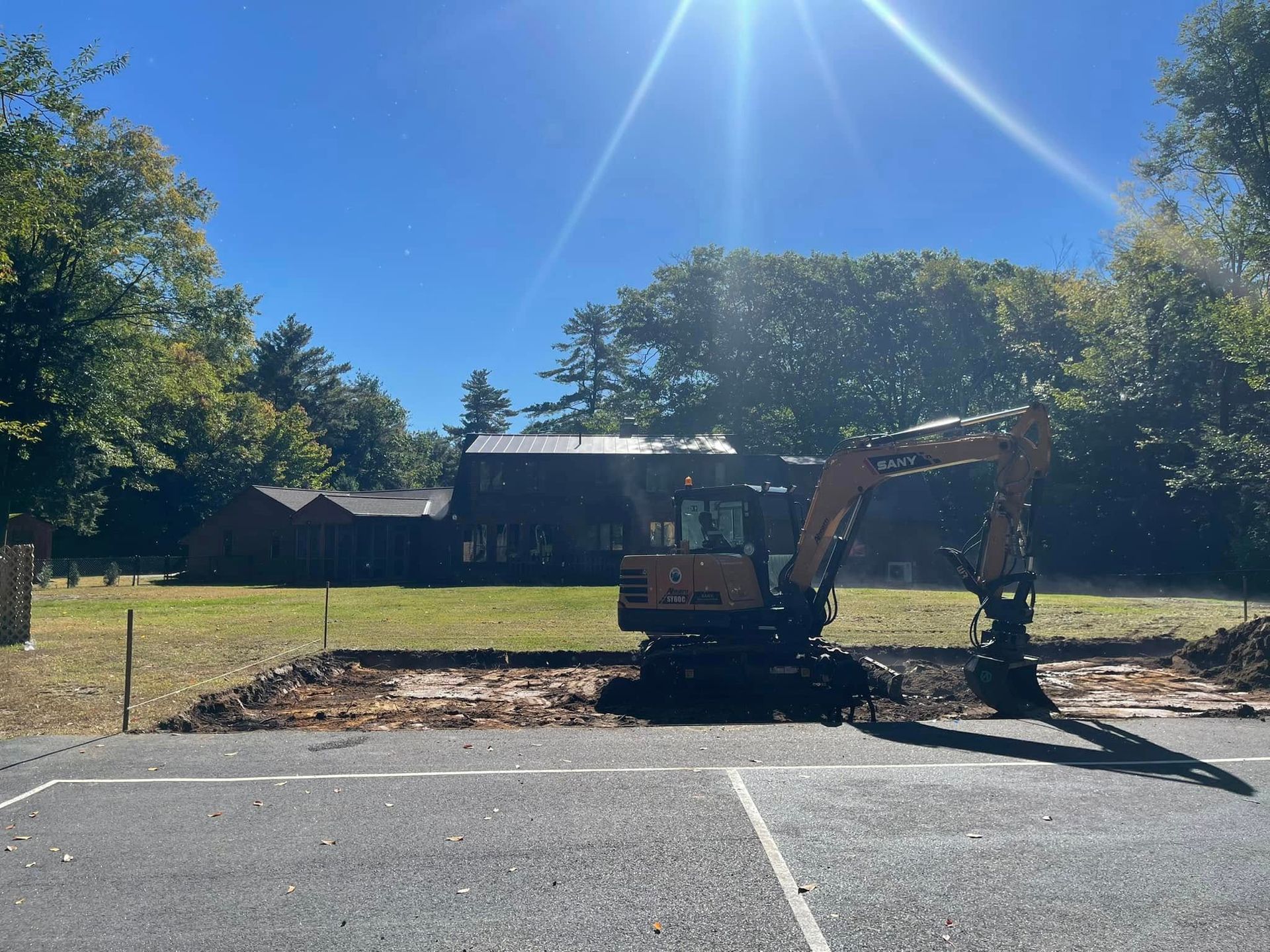 Excavator digging in an asphalt lot; trees and a building in the background on a sunny day.