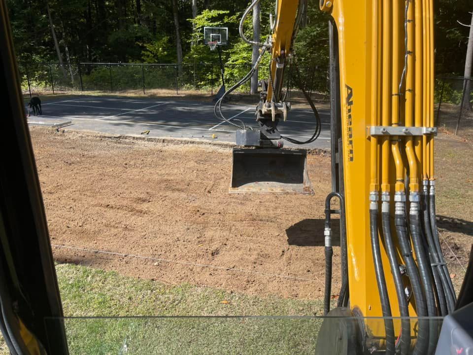 Excavator digging hole near basketball hoop on asphalt. Yellow machine, brown soil. Sunny day.