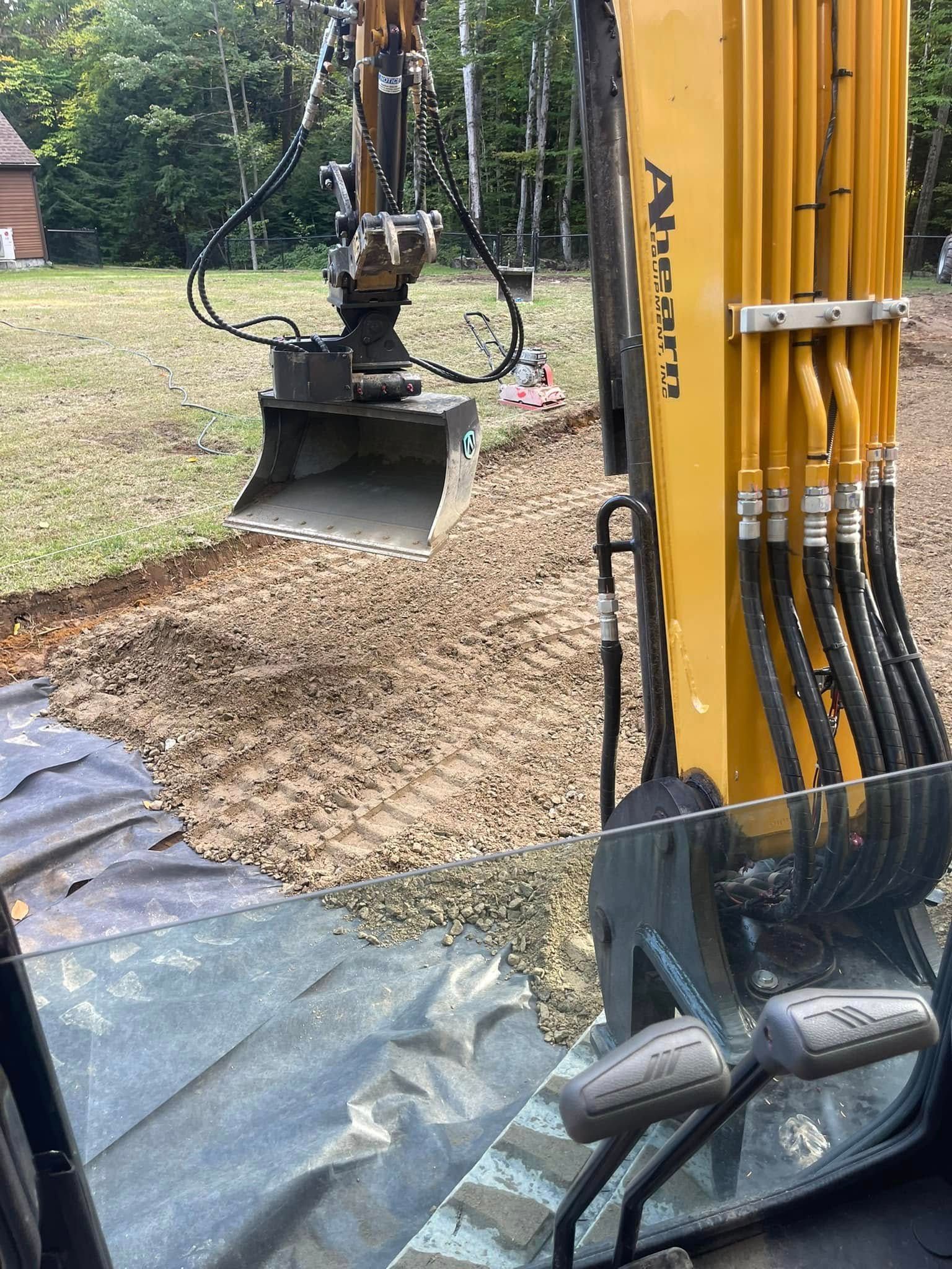Excavator digging a trench in a yard, visible cab controls, gravel and earth present.