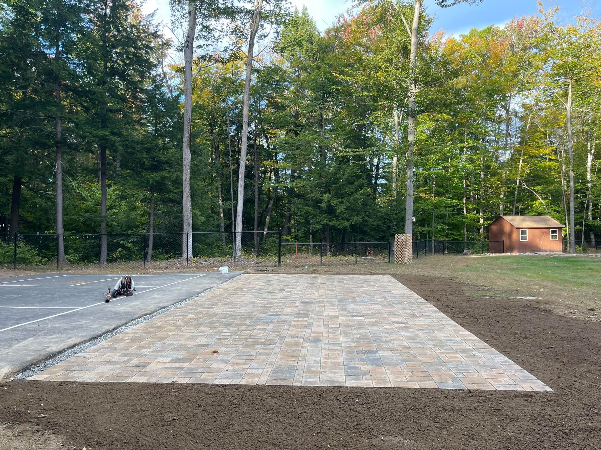 Paved patio with landscaping next to an asphalt area; small shed in the background.