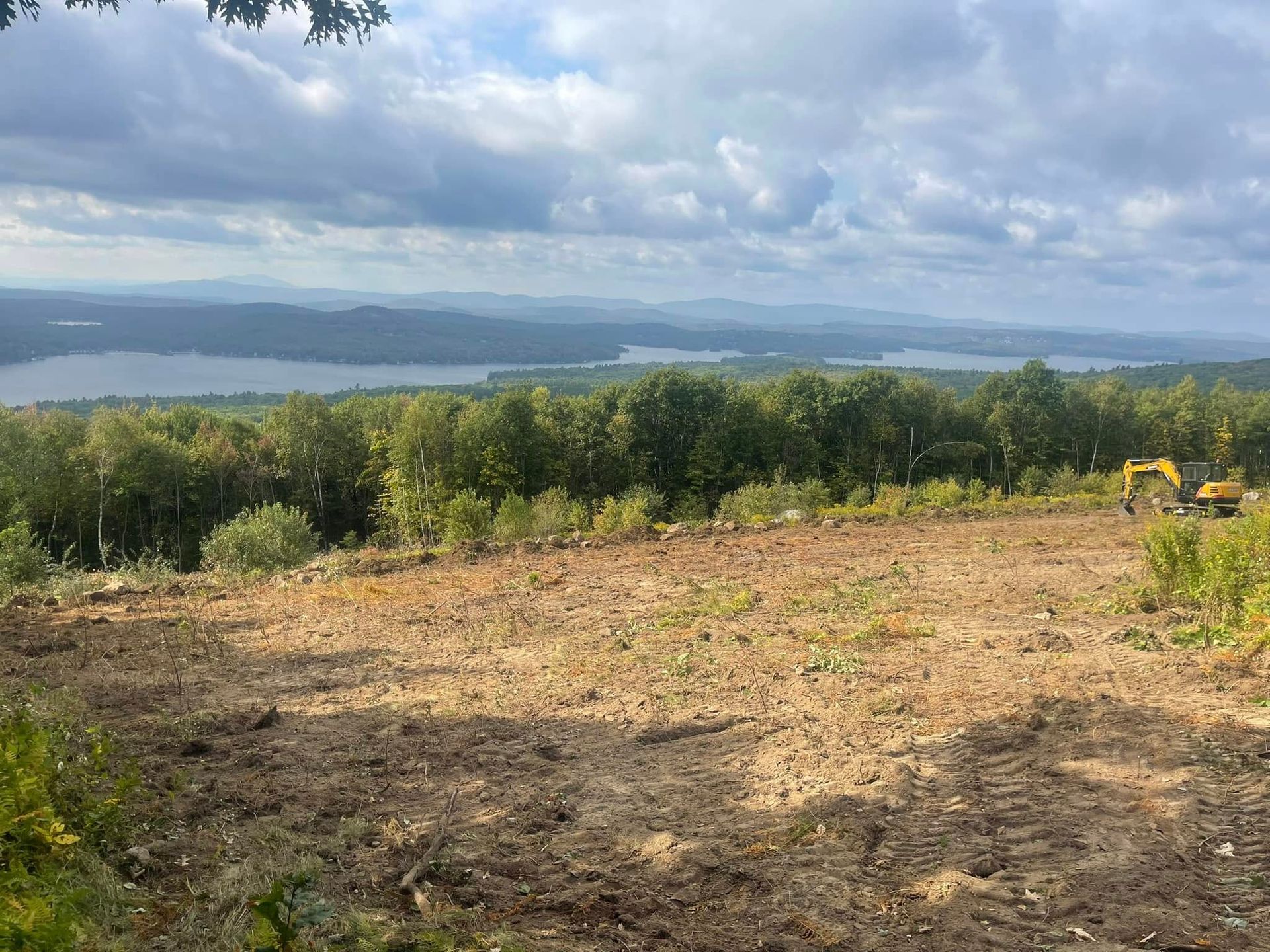 Cleared hilltop overlooking a lake and distant mountains; excavator visible on the right.