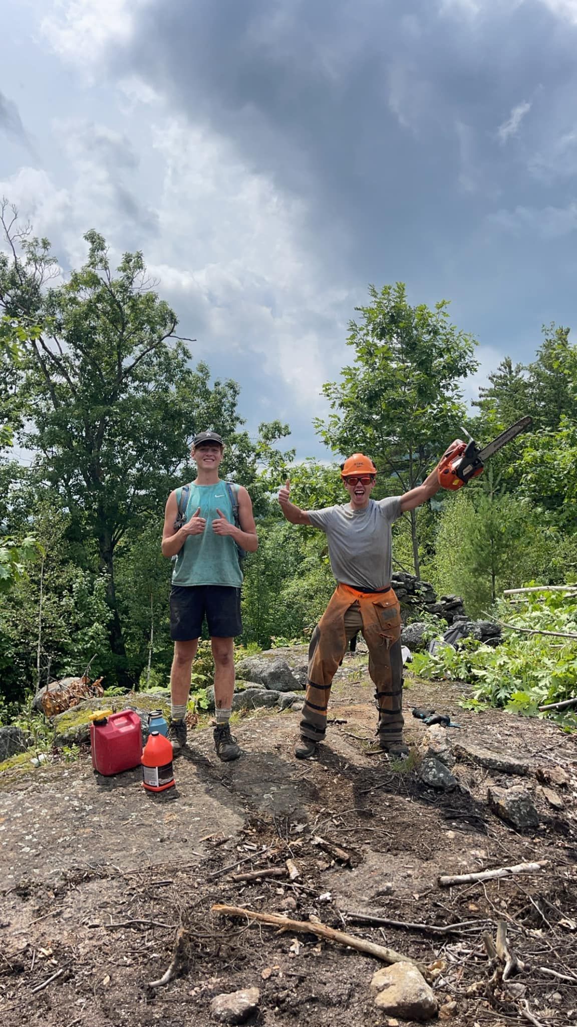 Two people on rocky ground. One holds a chainsaw, gesturing. Other with water bottles. Green trees and sky.