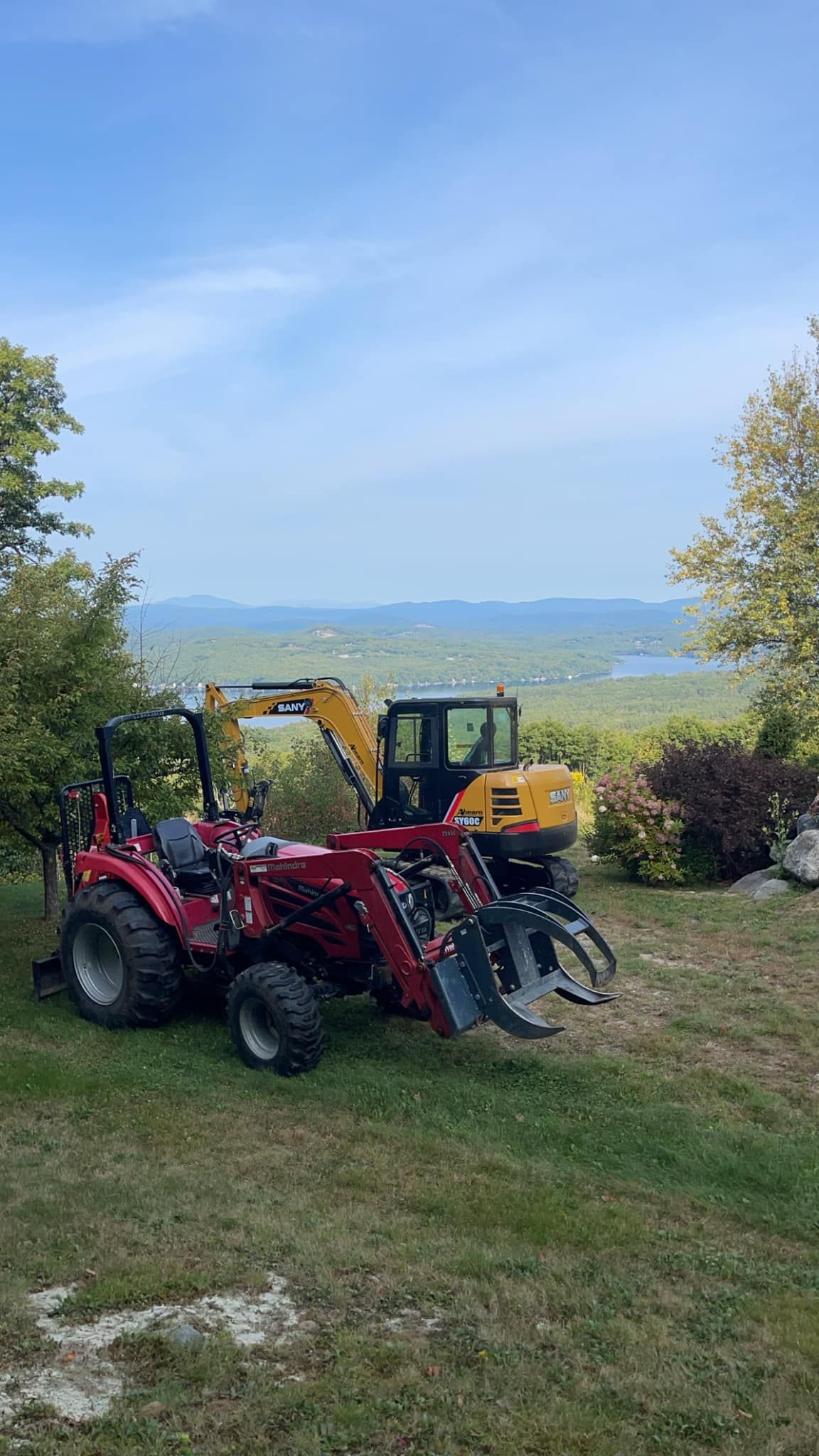 Red tractor with grapple and yellow excavator overlooking a scenic vista.