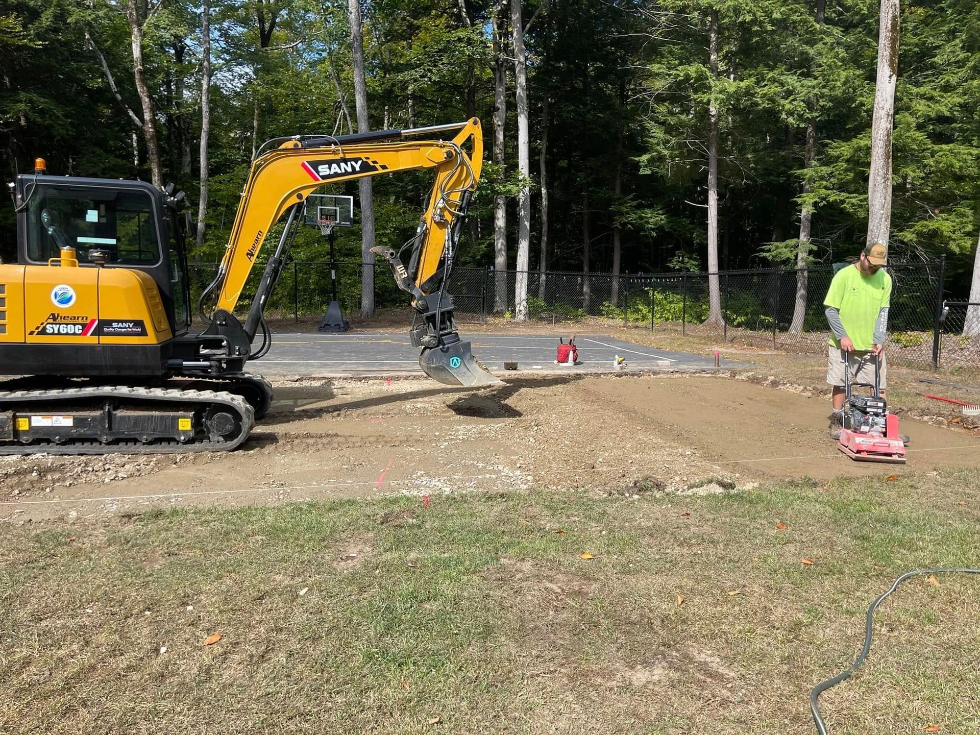 Construction site: Excavator and worker compacting soil with a vibratory plate compactor.