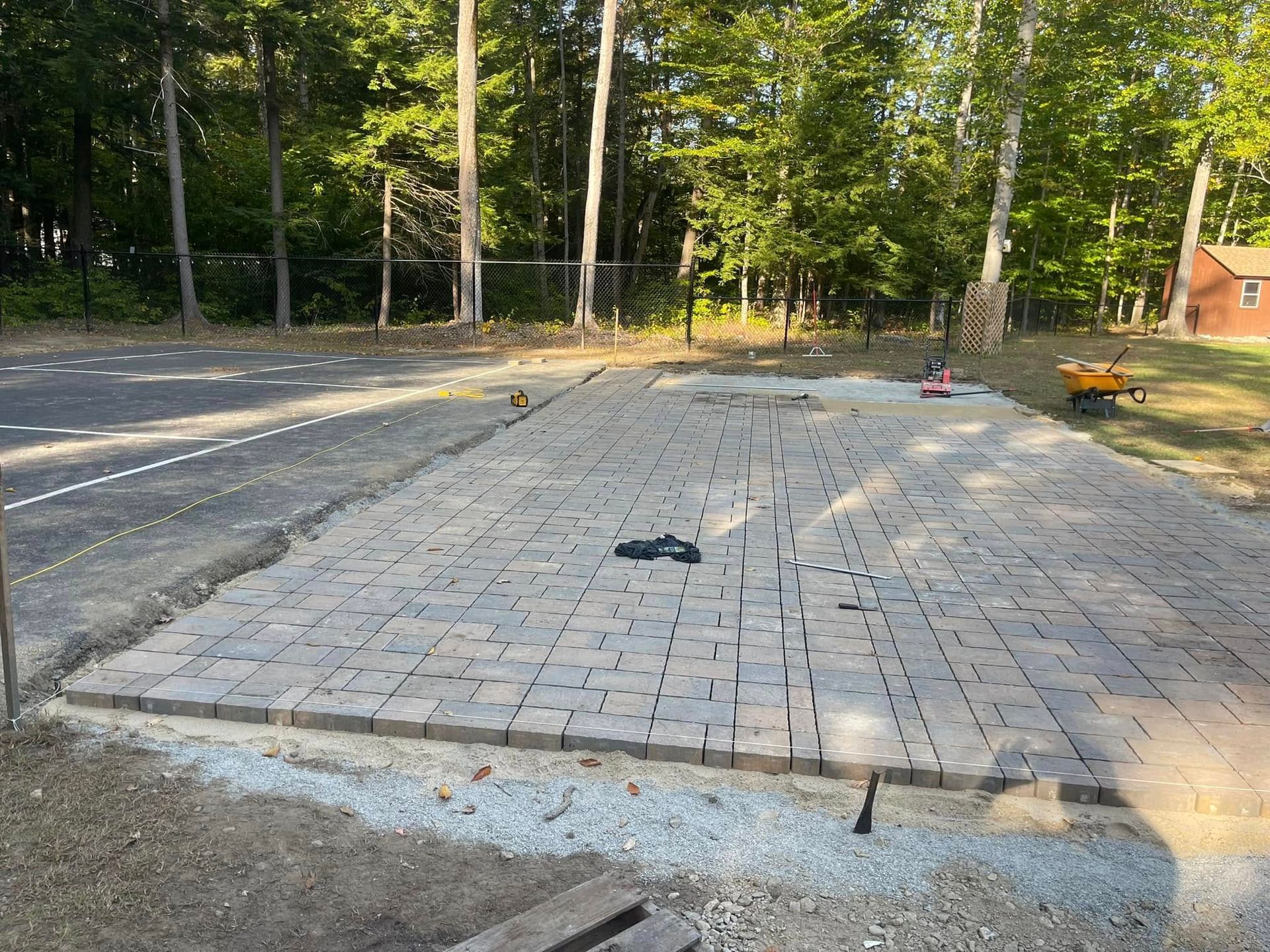 Pavers laid for a patio area next to a paved driveway. Tools, trees, and a house are in the background.