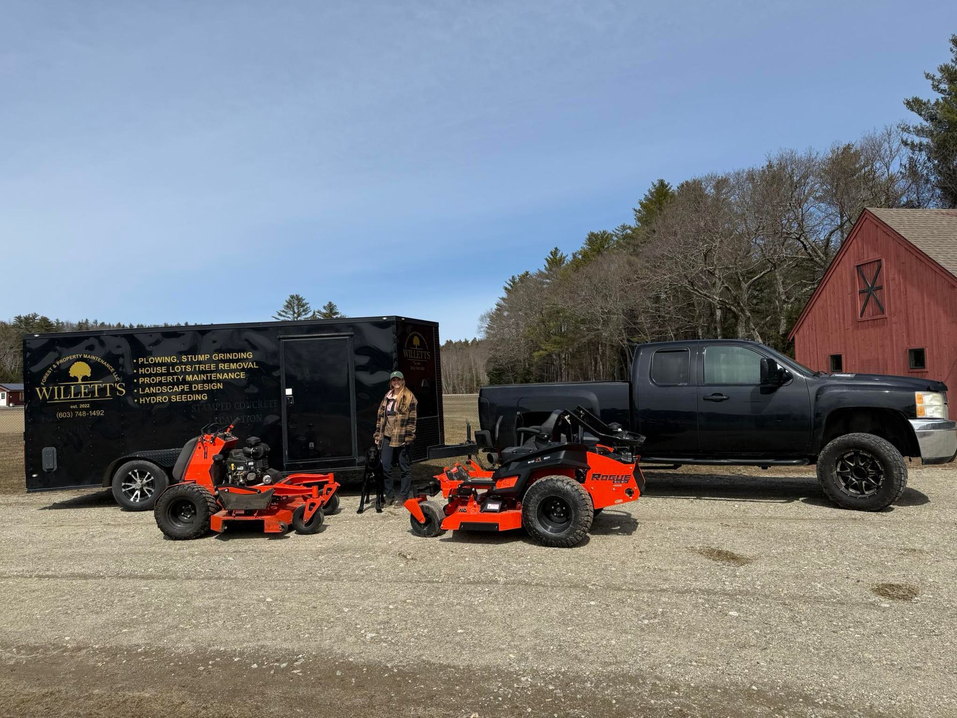 A black pickup truck, trailer, and two orange zero-turn mowers parked near a red barn. A person stands between the mowers.
