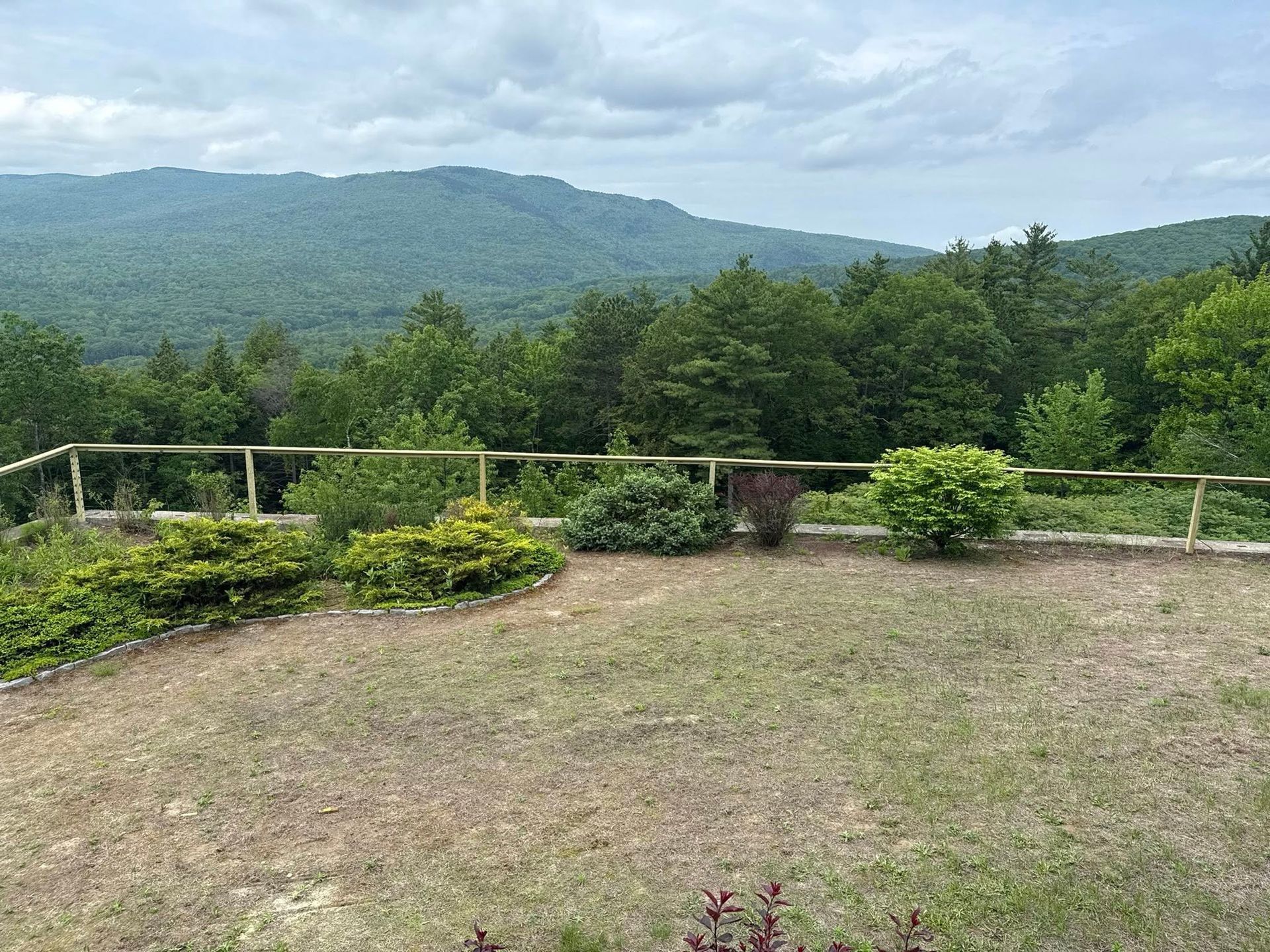 Grassy clearing with low fence overlooking a lush green forest and distant mountains under a cloudy sky.