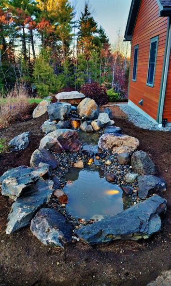 Stone-lined water feature under construction, with boulders and a small pond in a yard, trees in the background, next to a house.
