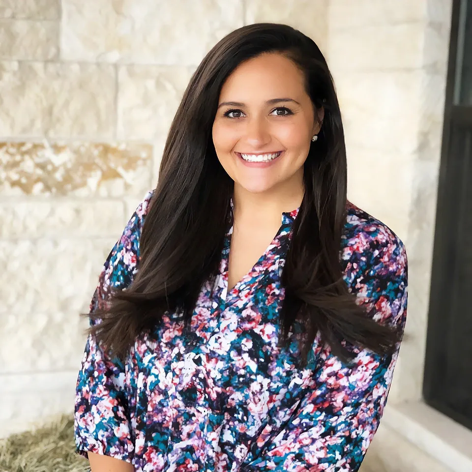 Smiling person in a floral blouse standing indoors against a light stone wall