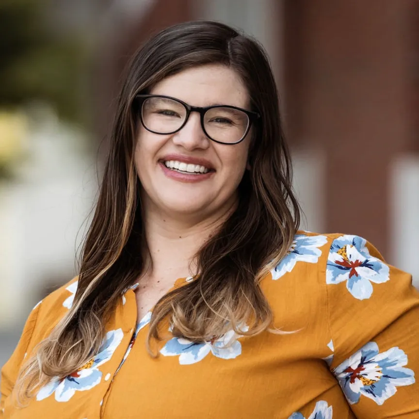 Smiling woman in black glasses and mustard floral blouse outdoors, with blurred brick background
