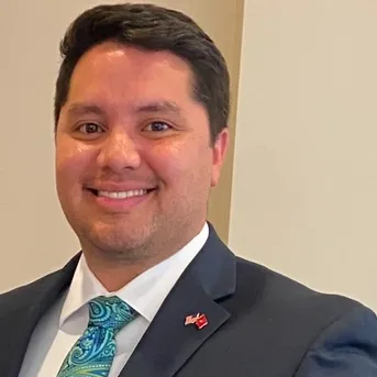 Smiling man in a dark suit and teal tie against a plain beige background