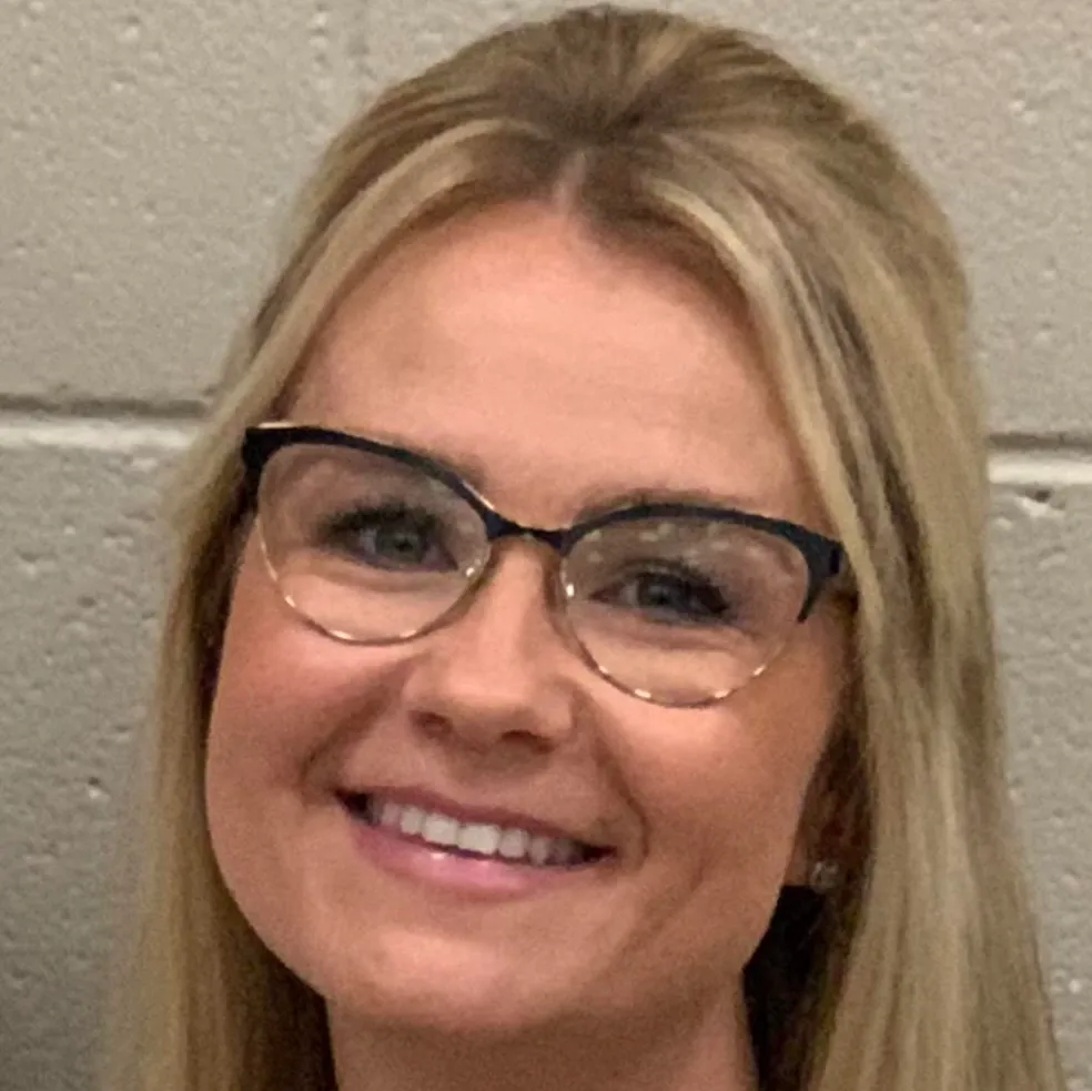 Smiling woman with blonde hair and glasses in front of a light gray wall