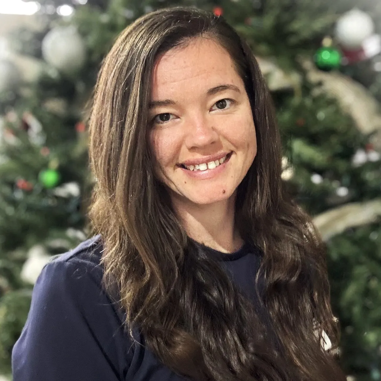 Smiling person with long brown hair in a navy shirt, standing in front of a blurred Christmas tree.