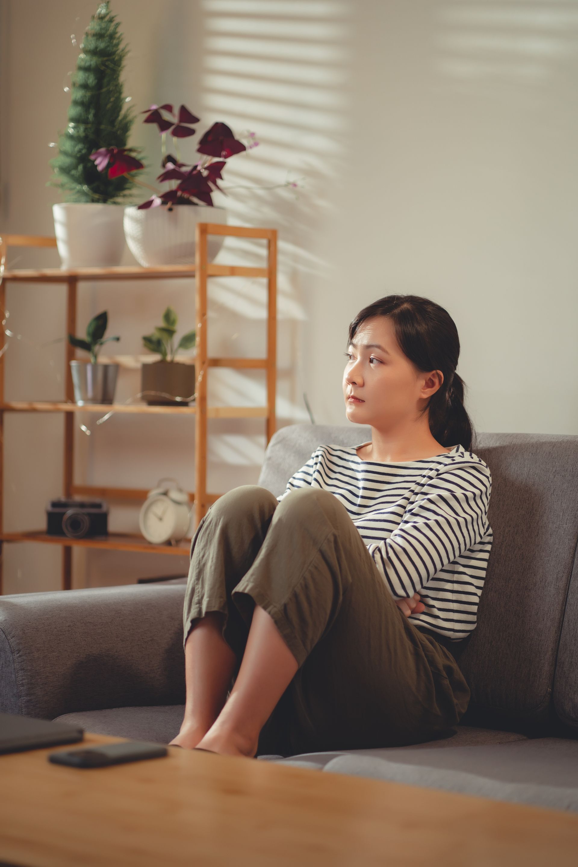Person sitting on a couch in a sunlit living room, looking out the window, with plants and shelves nearby.