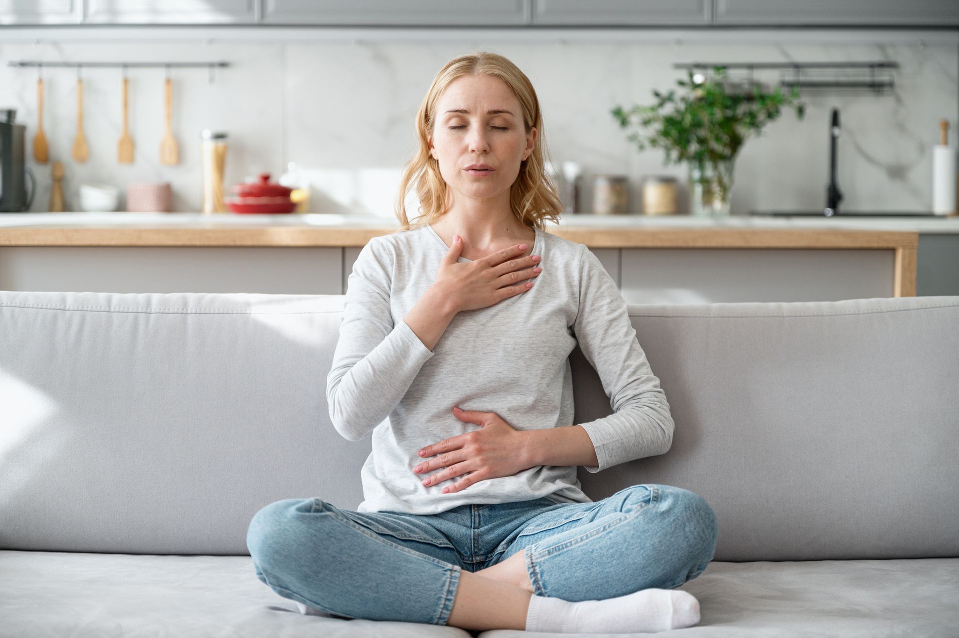 Woman sits on a sofa, holding her throat and stomach, appearing unwell in a bright living room.