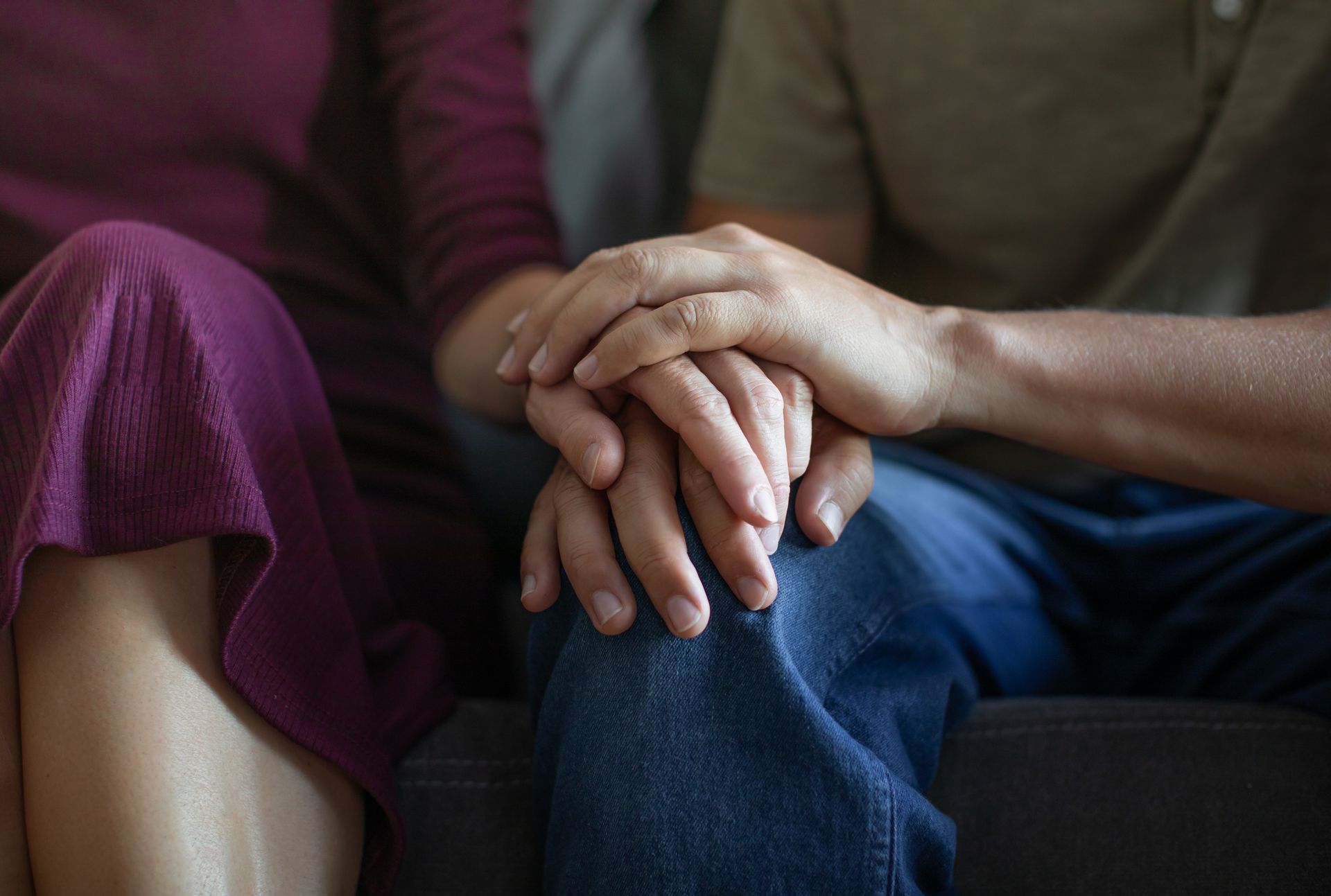 Two people sit close on a couch, holding hands in a comforting gesture.