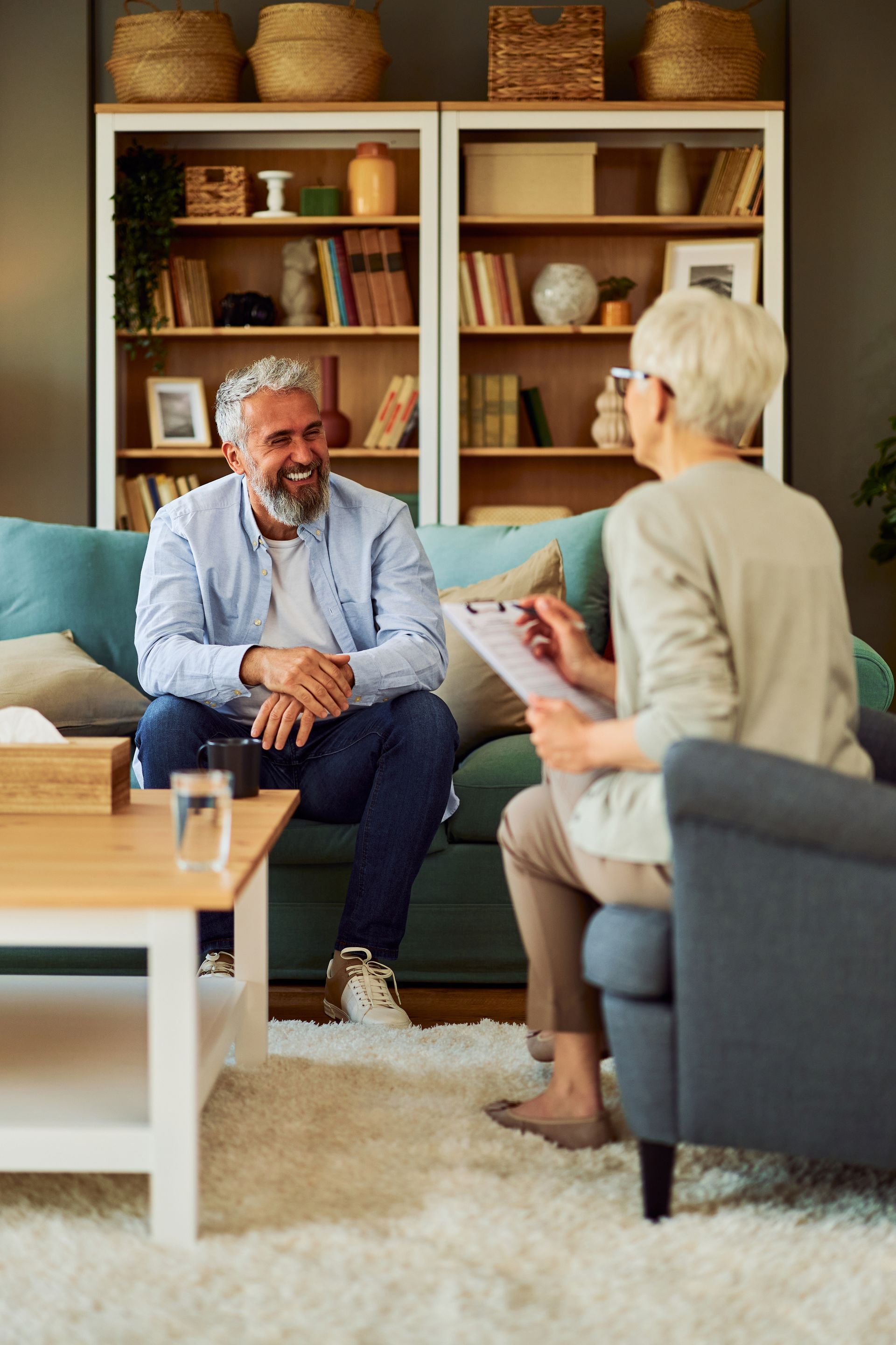 Two people talking in a cozy living room with bookshelves, a sofa, and a coffee table.