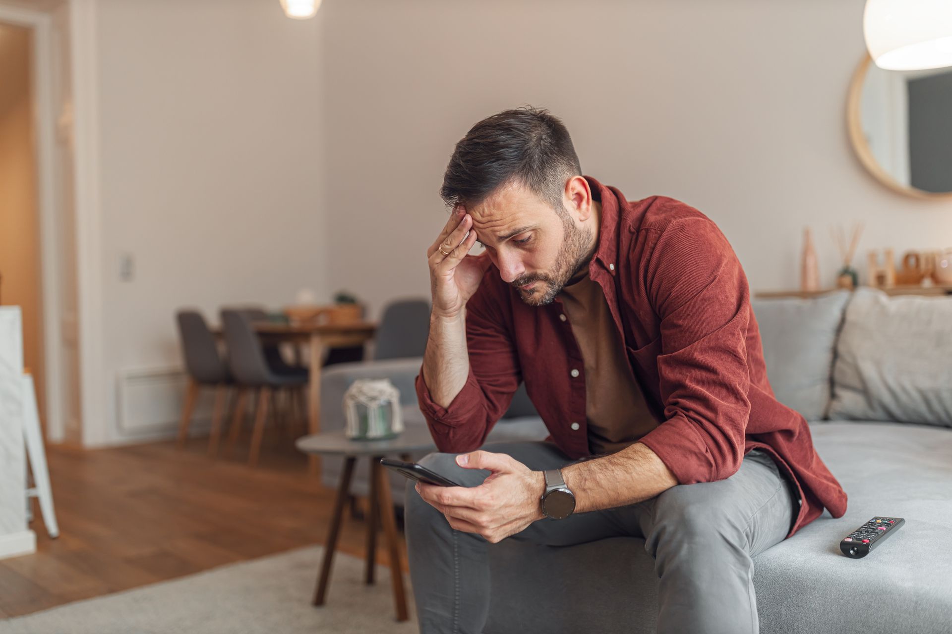 Man sitting on a couch, looking stressed while holding his head and checking his phone in a living room.