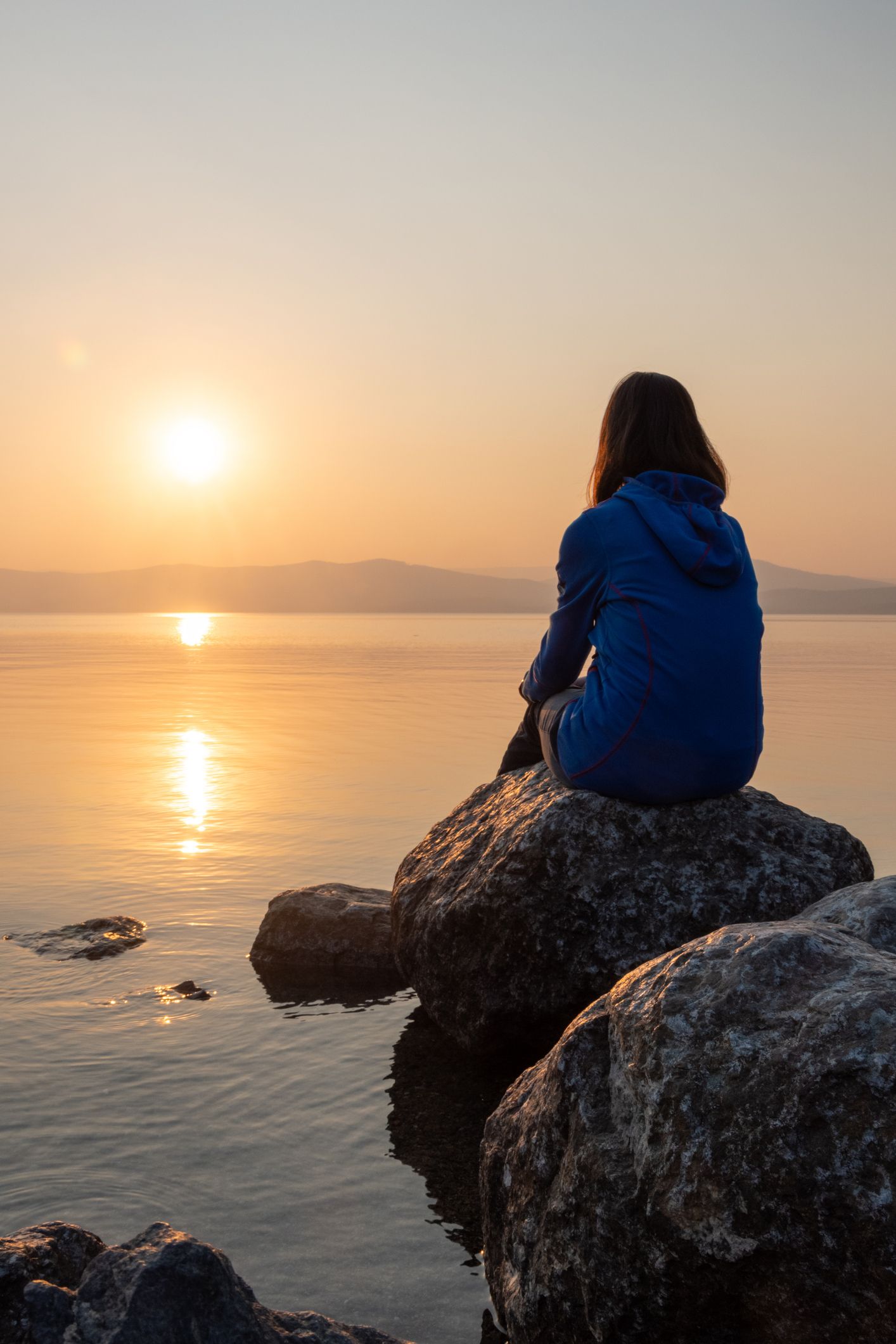 Person in blue jacket sitting on rocks at sunset, looking over calm water.