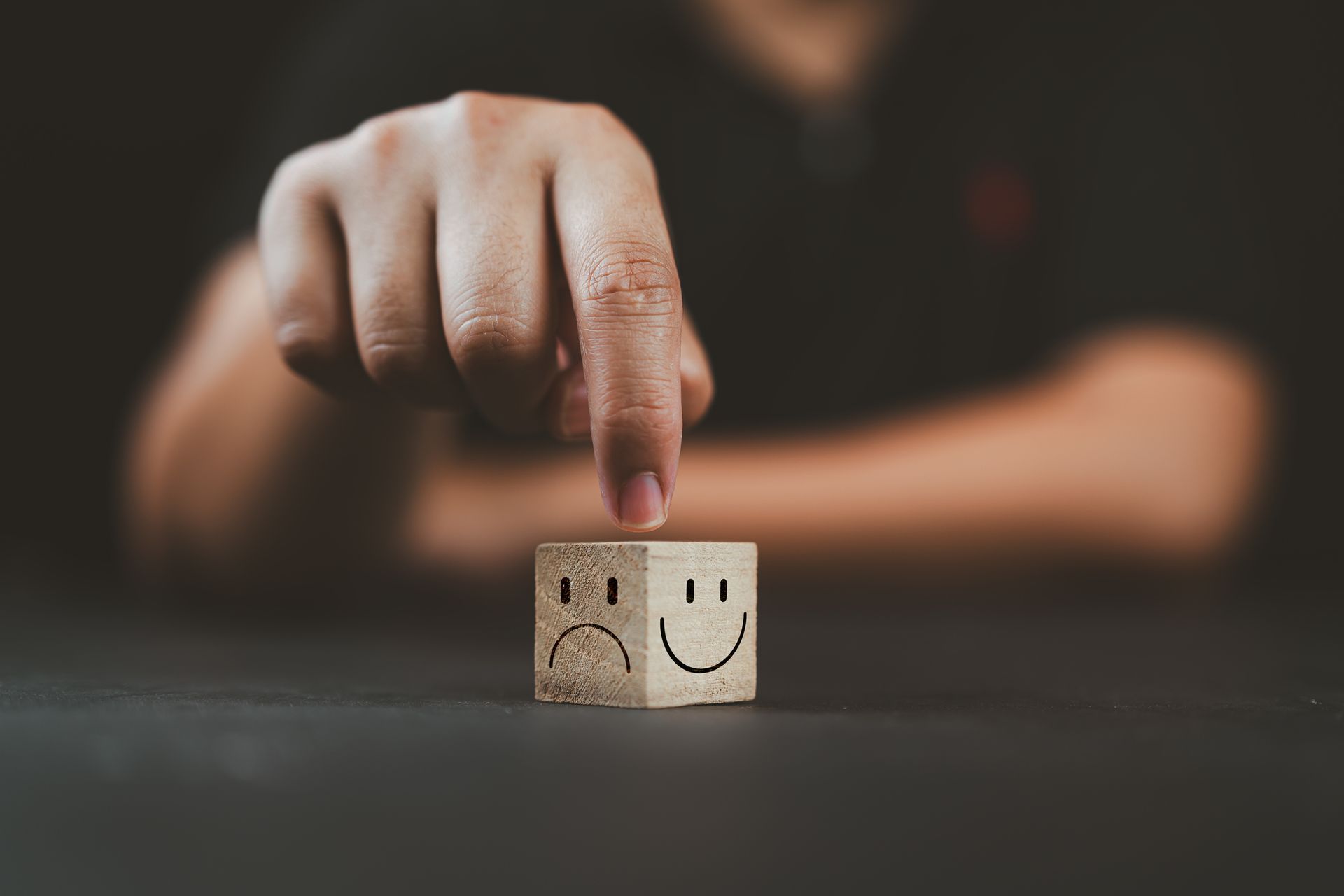 Hand points to two wooden blocks with sad and happy faces on a dark table.