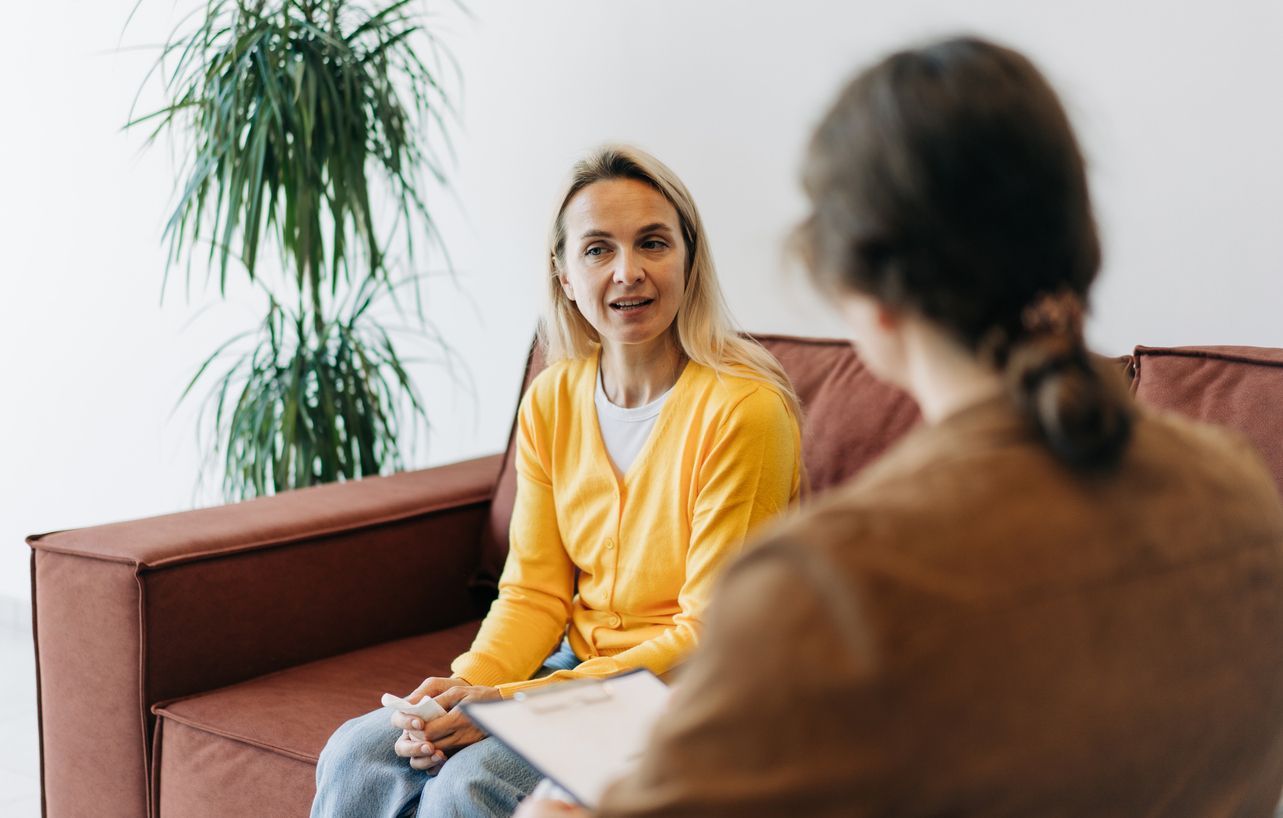 Two people talking on a sofa in a bright room with a potted plant