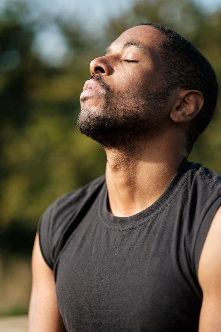 Man with eyes closed, basking in sunlight outdoors, wearing a black sleeveless shirt.