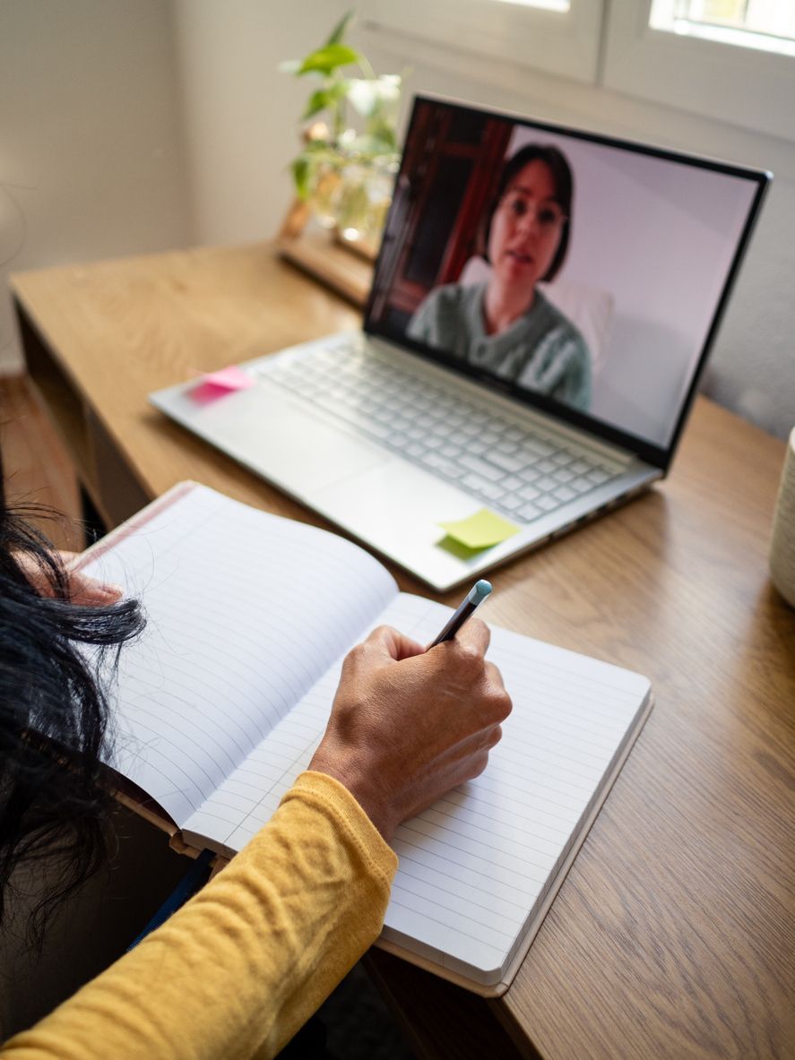 Person taking notes during a video call on a laptop at a wooden desk, with a notebook open in the foreground