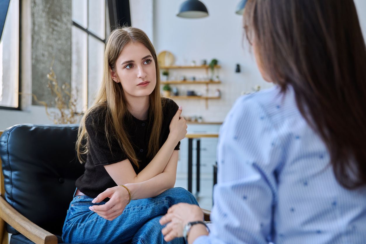 Two people talking in a bright room, one seated on a black couch and listening attentively.