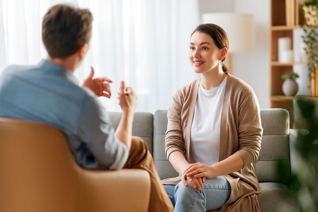Two people talking in a bright living room, one smiling on a sofa during a conversation.