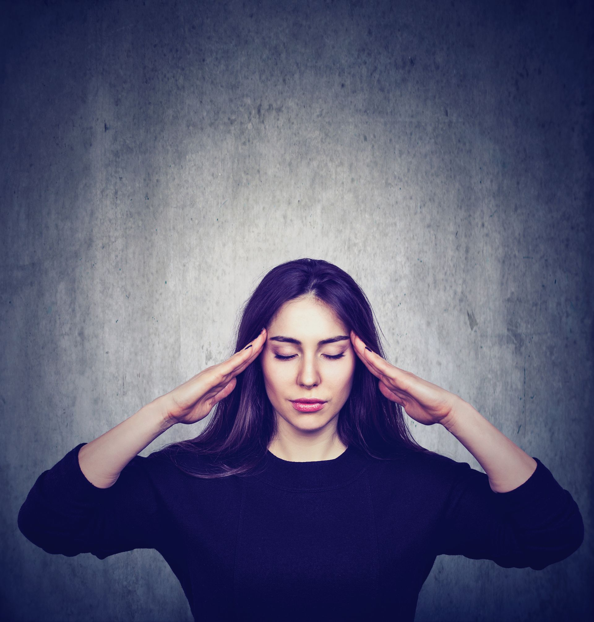 Woman in a dark sweater pressing her temples against a gray textured wall, eyes closed.