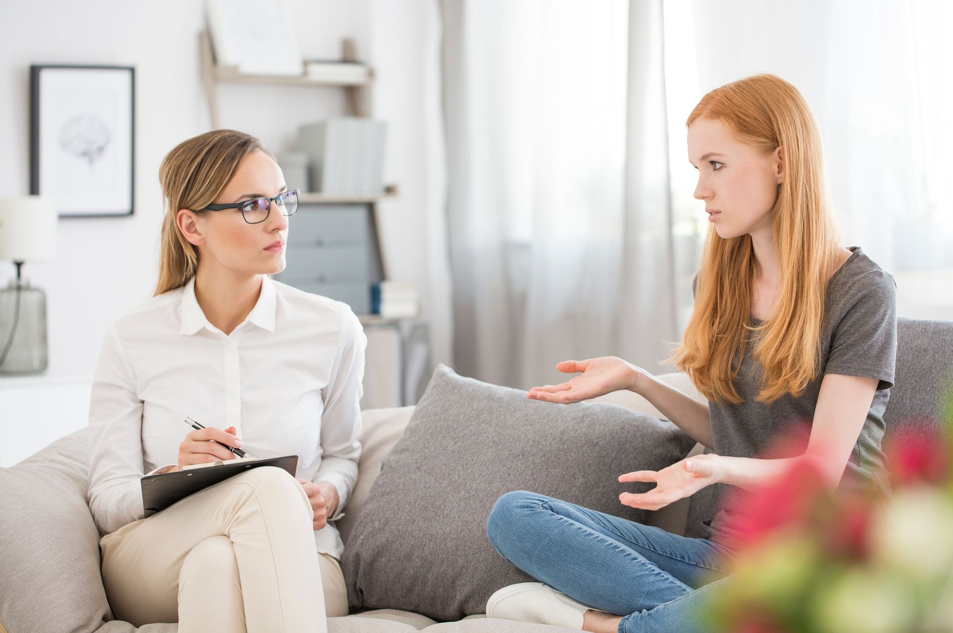 Two women talking on a couch in a bright living room, one holding a notebook while the other gestures.