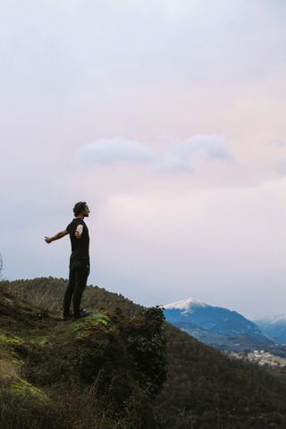 Person standing on a grassy cliff with arms outstretched, overlooking distant mountains under a pastel sky