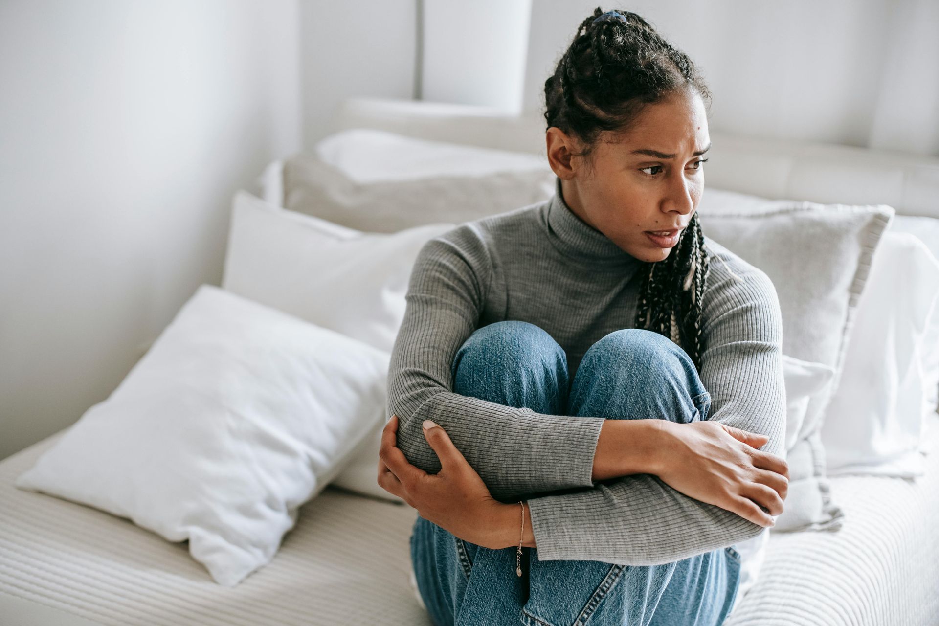 Person sitting curled up on a bed, hugging knees, looking away in a bright bedroom