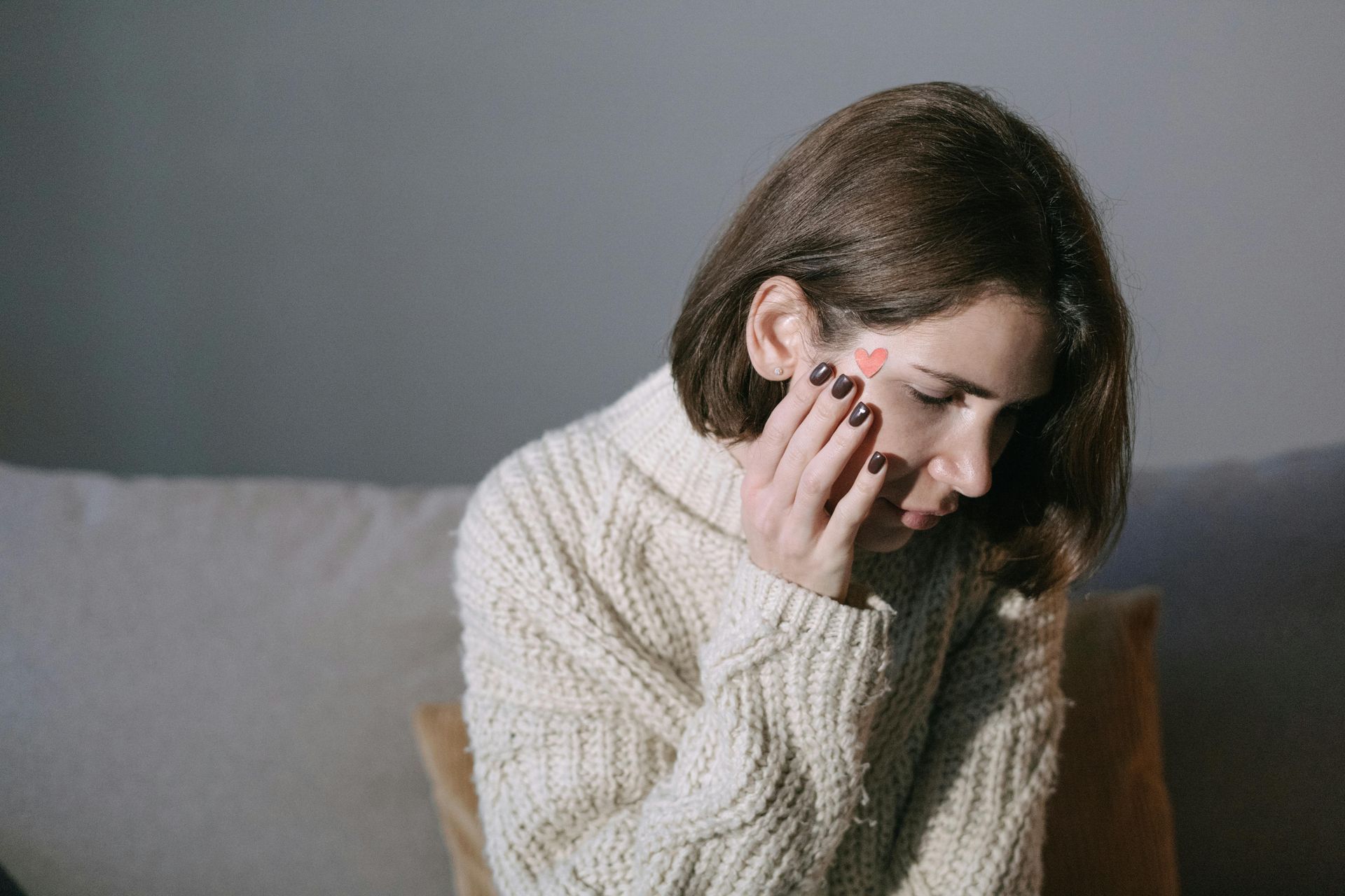 Person sitting on a couch, holding their face and looking down, wearing a cream sweater.