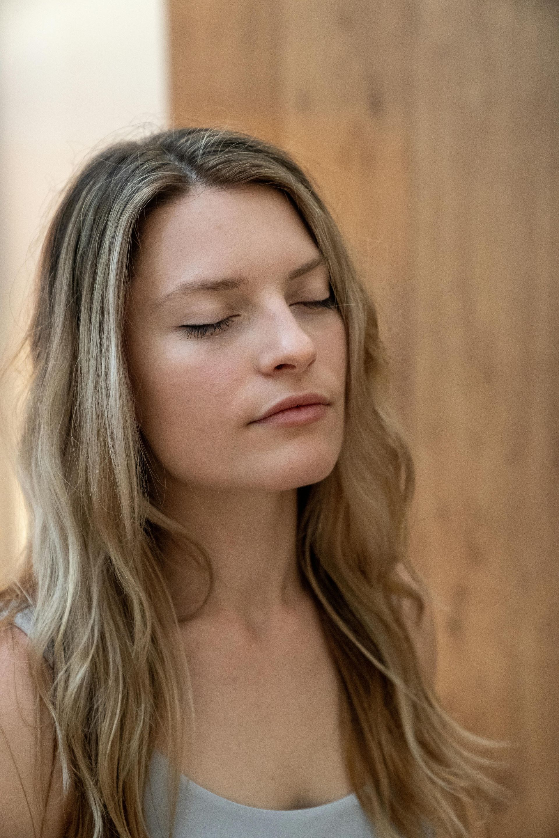 Woman with closed eyes standing against a wooden wall, wearing a gray top.