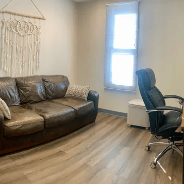 Small office with a brown leather sofa, desk chair, and two windows on light wood flooring.