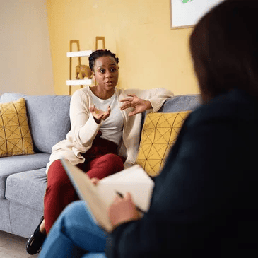 Two people in a counseling session, one speaking while seated on a gray couch with yellow pillows.