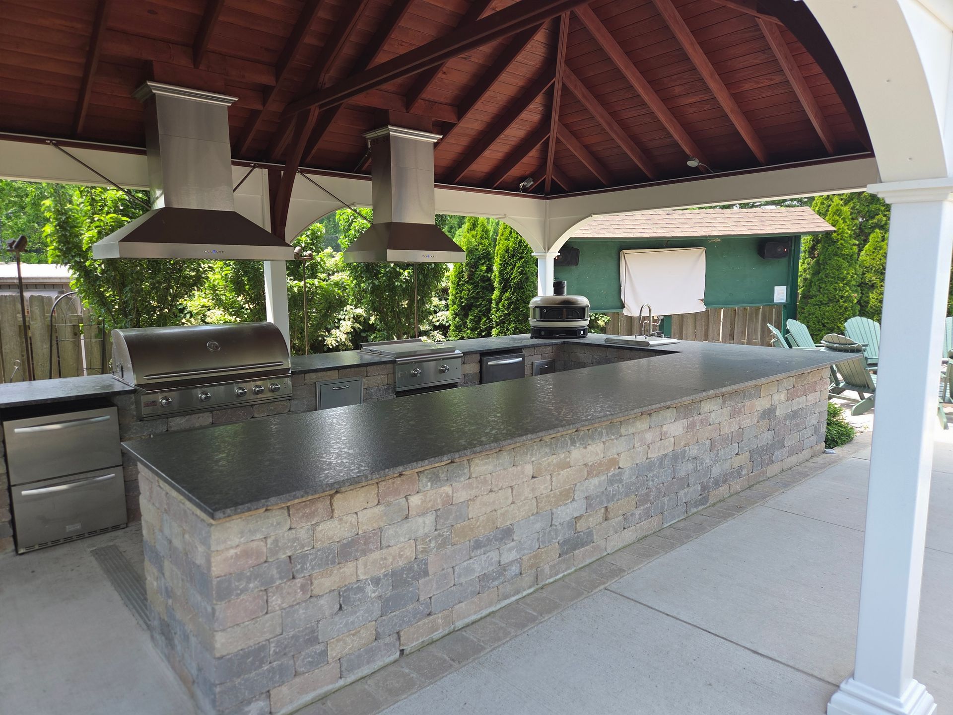 Outdoor covered kitchen with stone island countertop, built-in grill, and hanging stainless steel vents
