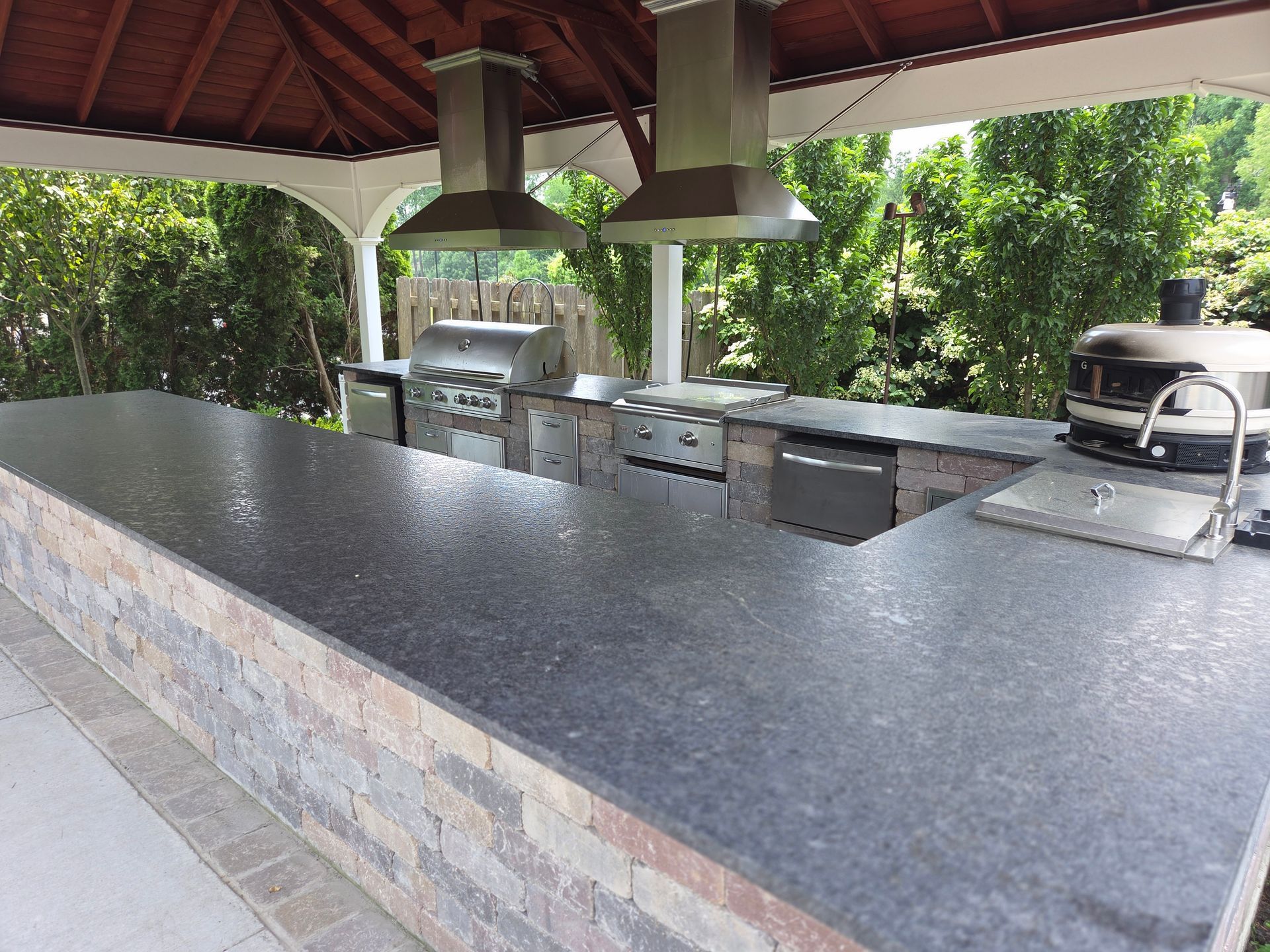 Outdoor covered kitchen with a long stone counter, grill, and metal hood under a red roof.