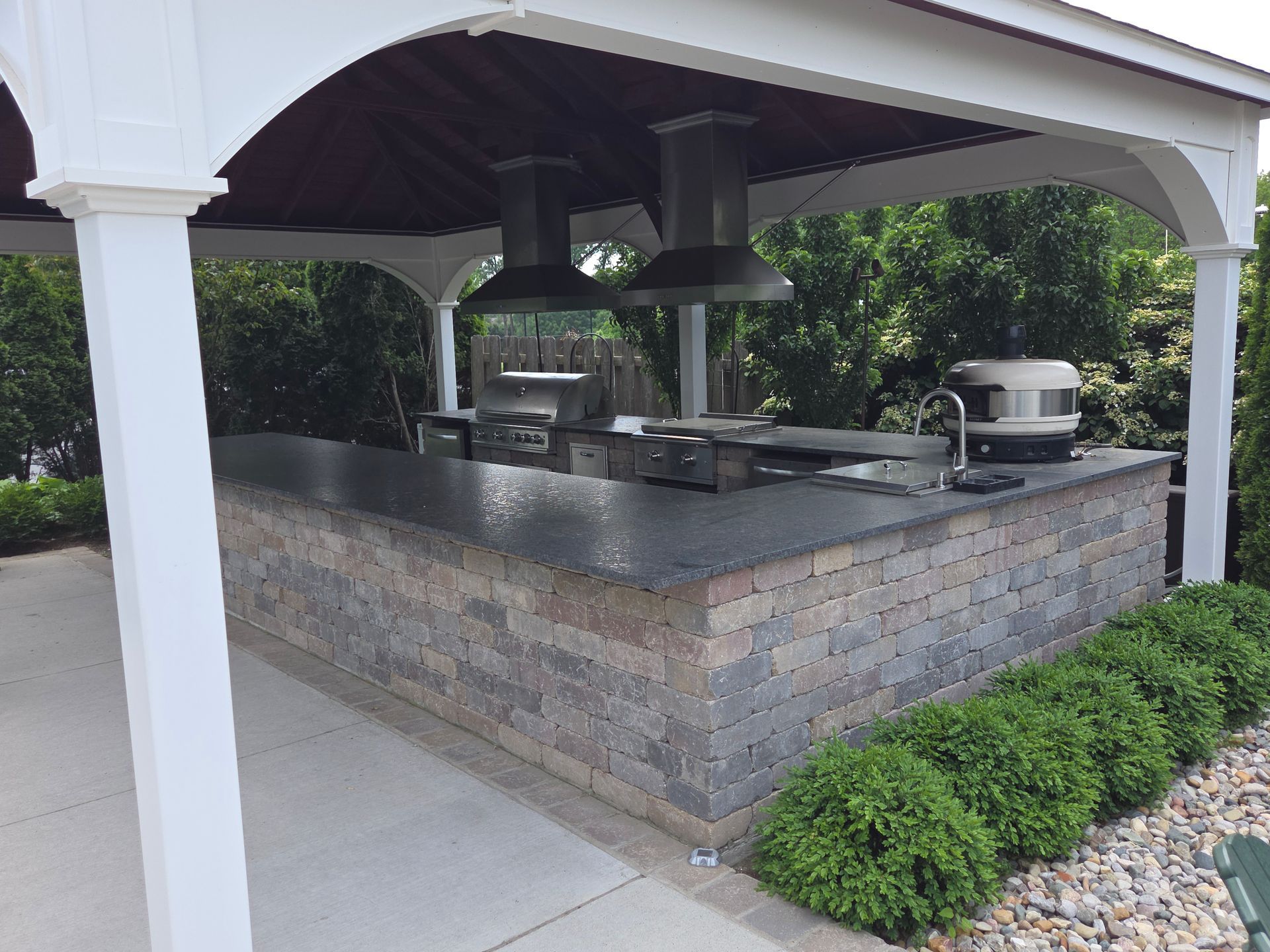 Outdoor covered kitchen with stone counters, built-in grill, and stainless steel vent hoods under a pavilion